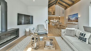 A modern living room with a fireplace, a sofa with a 'SKI JUDGE' pillow, a coffee table with books and a lantern, and a dining area in the background.