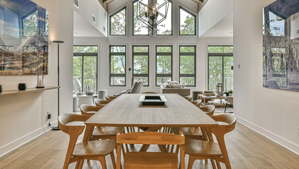 A spacious dining area with a long wooden table and chairs, large windows, and a high ceiling with exposed beams.