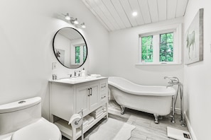 A modern bathroom with a freestanding bathtub, a round mirror, and a window with a view of greenery. - La Maison Blanche | Hot Tub & Sauna | Pool Table, Fire Pit & Close to Resort (Mont-Tremblant)