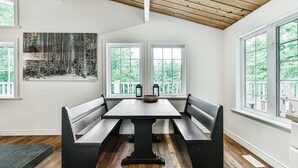 Un comedor con mesa y bancos de madera, ventanas amplias con vista a árboles, y un cuadro en la pared.