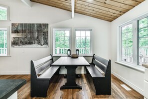 Ein Essbereich mit Holztisch und Bänken, großen Fenstern mit Blick auf Bäume und einem Gemälde an der Wand.