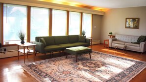 Een woonkamer met een groene bank, een witte bank, een kamerkleed met patroon en grote ramen met zonwering.