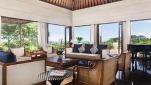 A spacious living area with a sofa, chairs, a coffee table, and a piano. Large windows offer a view of greenery and the ocean.