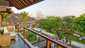 A spacious balcony with a wooden railing, a seating area with cushions, and a view of a lush garden with palm trees and a house in the background.