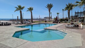 Un área de piscina con piscina rectangular, sillas de descanso y palmeras.