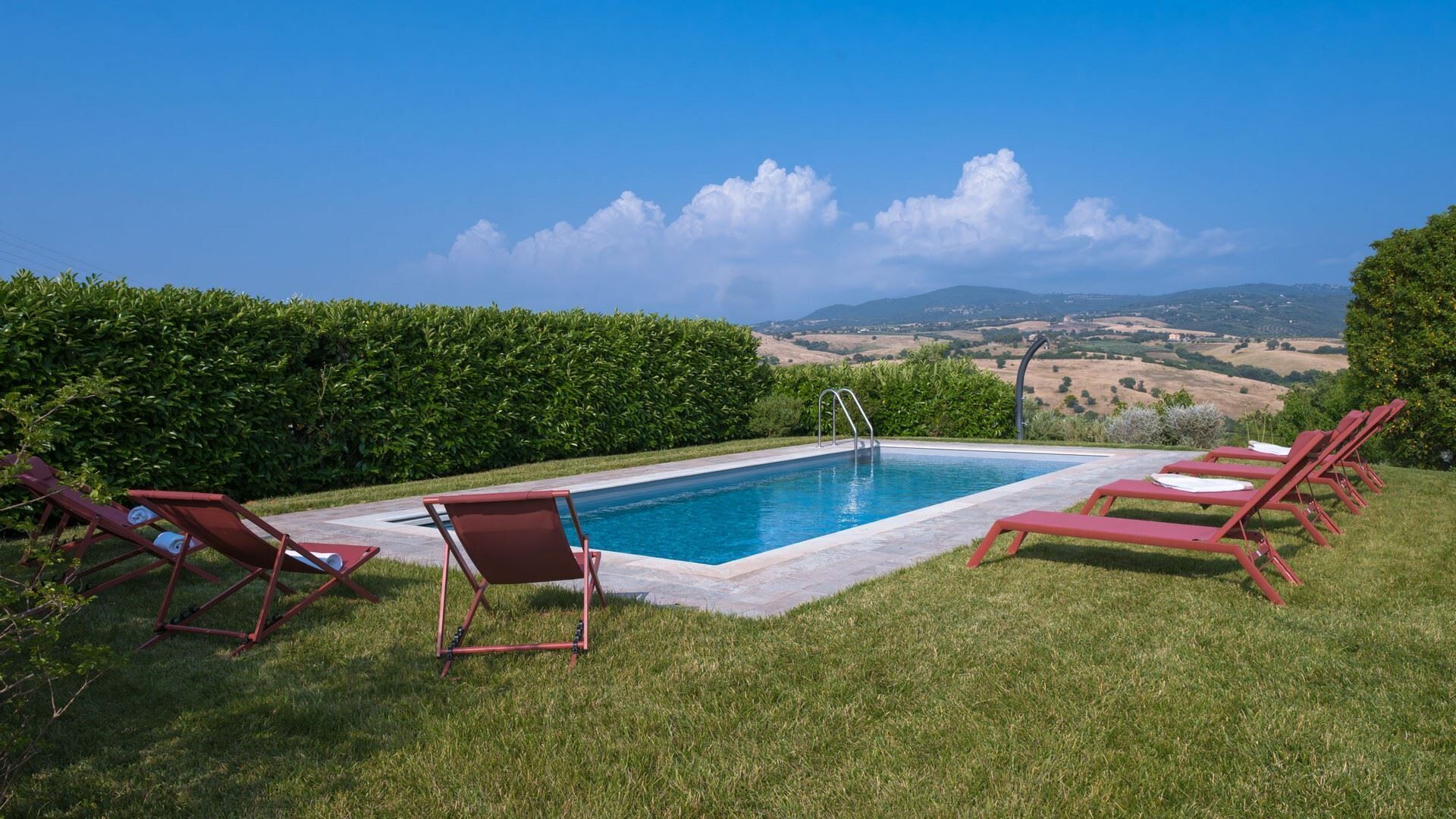 A rectangular swimming pool with a clear blue water surface, surrounded by a low stone wall. There are six red lounge chairs aligned in two rows on the grass, and a metal ladder leading into the pool. The background features a well-maintained hedge, a clear blue sky, and a distant view of rolling hills.