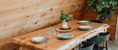 Une table à manger en bois, avec des assiettes et des verres, entourée de chaises noires, dans une pièce aux murs en bois et agrémentée d’une plante.