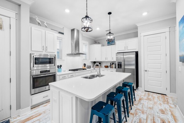 Una cocina moderna con armarios blancos, electrodomésticos de acero inoxidable, una isla central con taburetes y luminarias colgantes.
