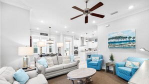 A spacious living room with a ceiling fan, a large painting, and a kitchen in the background.