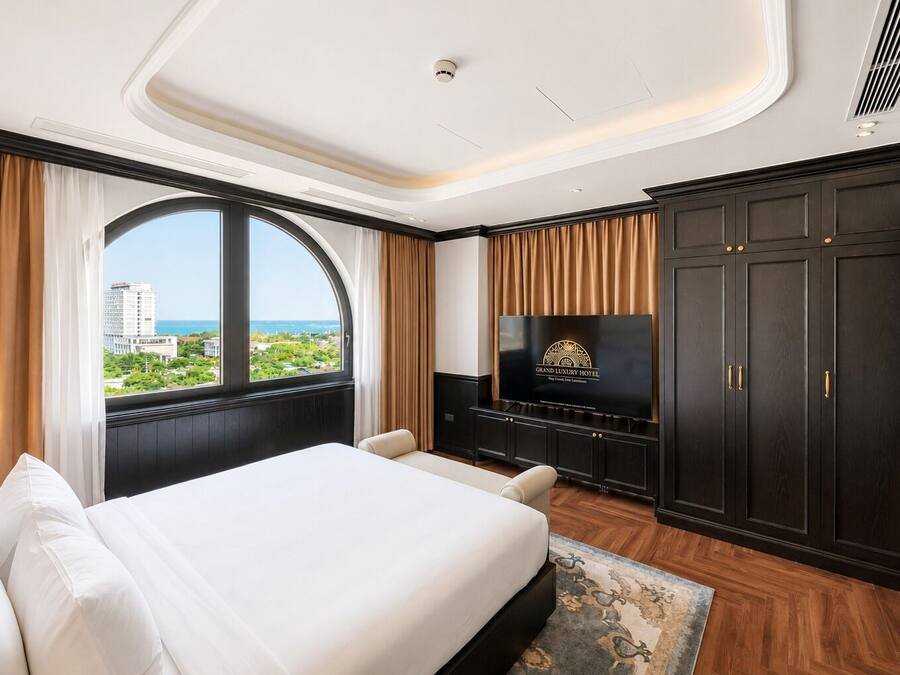 A hotel room with a large bed, a television, dark wood furniture, and a view of the city.