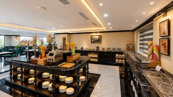 Un moderno vestíbulo de hotel con un mostrador de recepción de mármol, una zona de buffet con estaciones de comida y bebidas, y un área de descanso con sillas verdes.