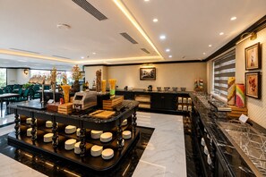 Un moderno vestíbulo de hotel con un mostrador de recepción de mármol, una zona de buffet con estaciones de comida y bebidas, y un área de descanso con sillas verdes.