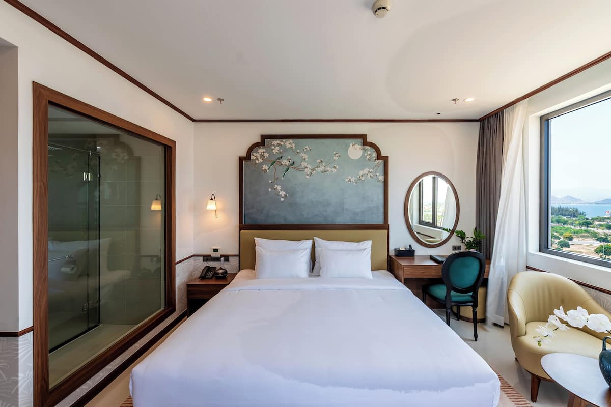 A hotel room with a large bed, a desk with a chair, a glass-enclosed shower, a view of the outdoors, and a decorative wall piece.