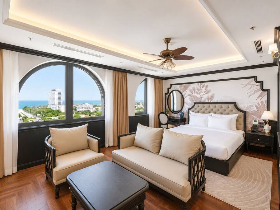 A hotel room with a large bed, two armchairs, a coffee table, a ceiling fan, and a view of the city.