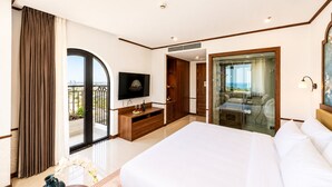 Habitación de hotel con una cama grande, televisión, balcón y zona de ducha con paredes de vidrio.