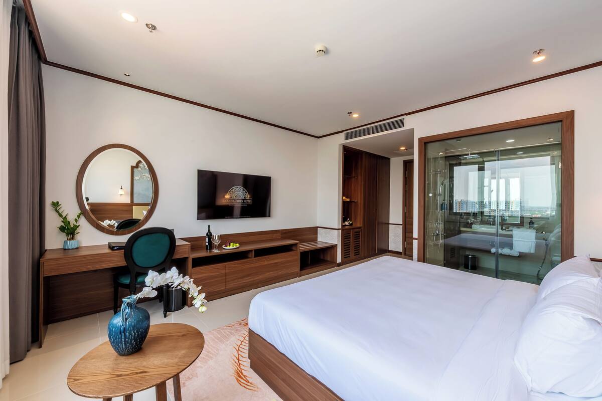 A modern hotel room with a large bed, a desk with a chair, a television, a mirror, and a bathroom with a glass shower.