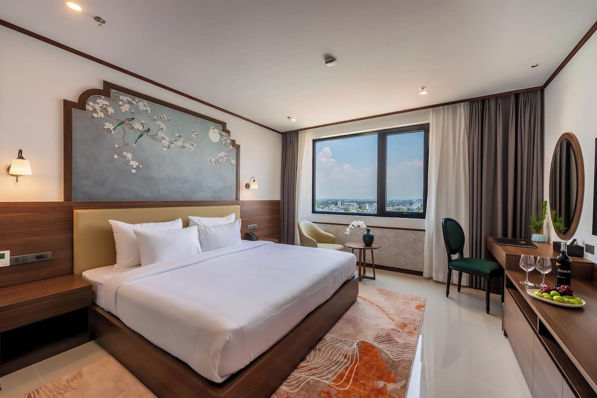A modern hotel room with a large bed, a desk with a chair, a small table with a vase, and a view of the city through large windows.