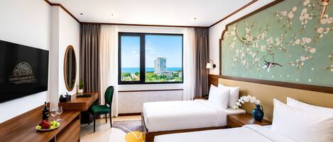 A hotel room with two beds, a desk with a chair, a television, and a large window with a view of a building and greenery.