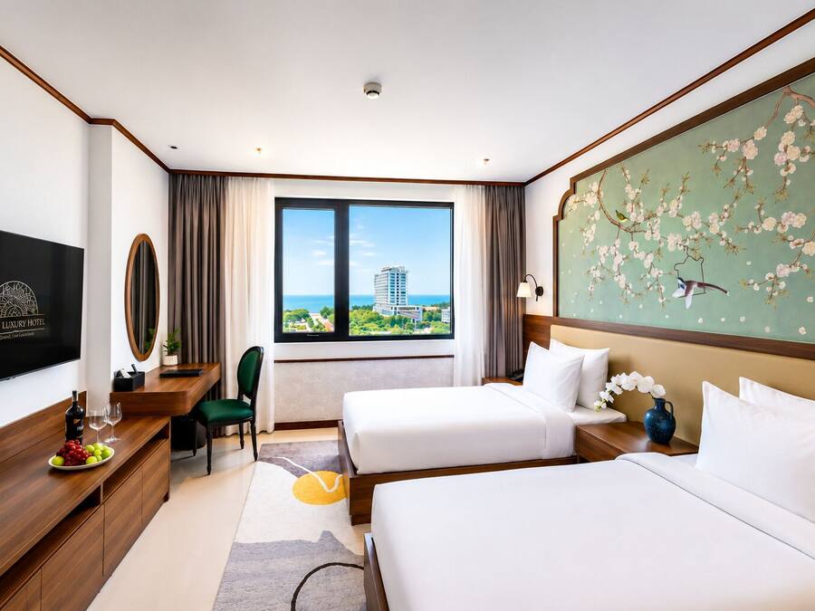 A hotel room with two beds, a desk with a chair, a television, and a large window with a view of a building and greenery.
