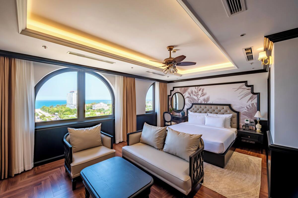 A hotel room with a large window, a bed, a sofa, a small table, and a ceiling fan.