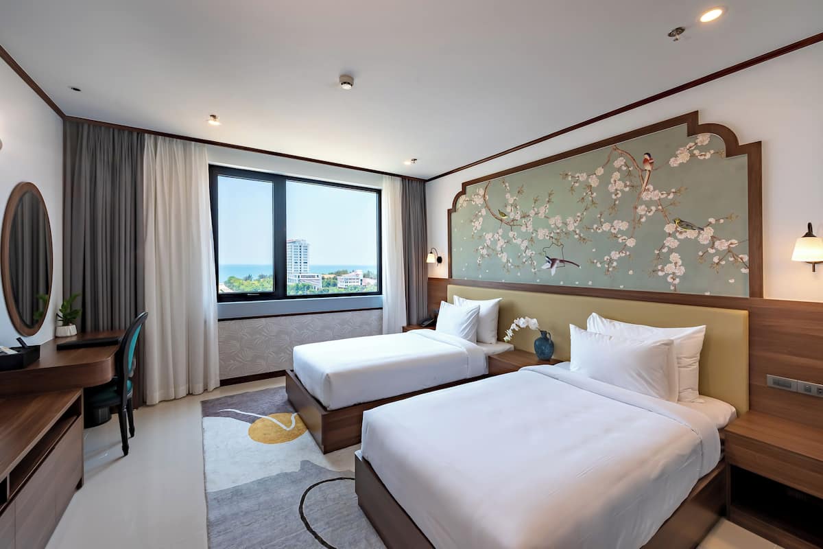A hotel room with two beds, a desk, a chair, a large window with a view, and a decorative wall piece.