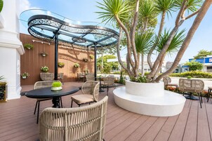 Un patio al aire libre con mesas redondas y sillas de mimbre, rodeado de plantas en macetas y una estructura metálica decorativa.