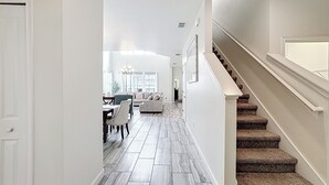 A hallway with a staircase leading to a second floor, a dining area with a table and chairs, and a living room with a sofa and a chandelier. - 🏝️ 5BR/5BA Home w/ Private Pool & Spa, Themed Kids’ Rooms & Resort Fun | Storey Lake Near Disney (Kissimmee)