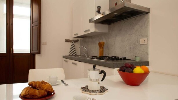 Eine Küche mit weißer Arbeitsplatte, einer Kaffeemaschine, einer Obstschale und einem Croissant auf dem Tisch.