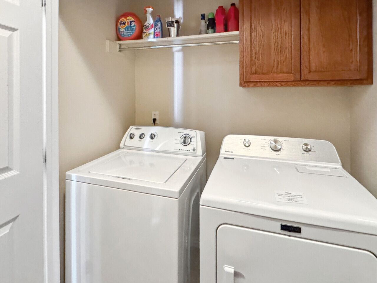 A laundry area with two white washing machines, a shelf with cleaning products, and wooden cabinets.