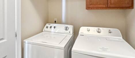 A laundry area with two white washing machines, a shelf with cleaning products, and wooden cabinets.
