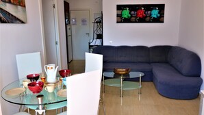 A living room with a glass dining table, white chairs, a blue sofa, and a wall-mounted picture of scooters.