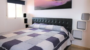 A bedroom with a large bed, a window with a dark blind, a wall-mounted artwork of a sunset, and a bedside table with a lamp.