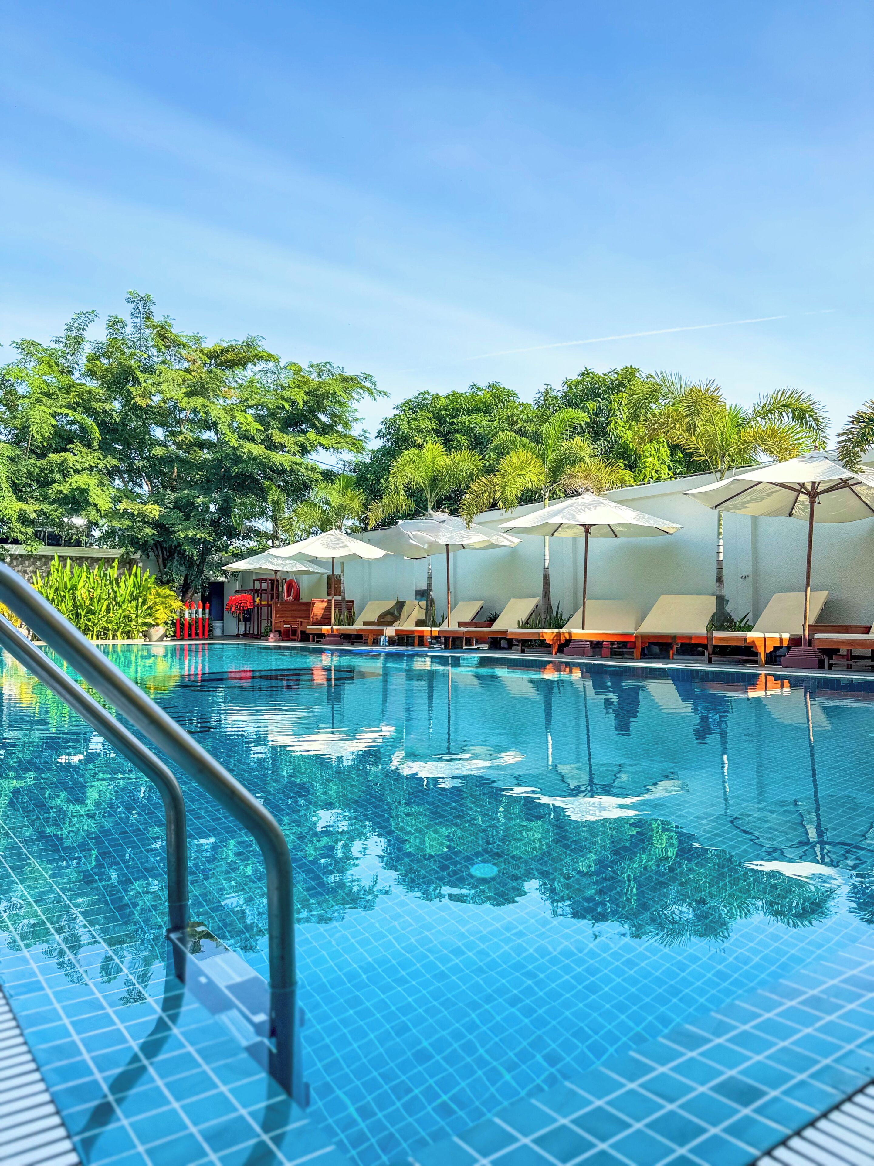 A swimming pool with clear blue water, surrounded by lounge chairs and umbrellas, and trees in the background.