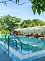 Una piscina con sillones de descanso y sombrillas, rodeada de árboles y un cielo despejado.