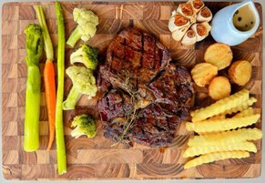 Una tabla de madera con un bistec, espárragos, brócoli, zanahorias, dientes de ajo y papas fritas.