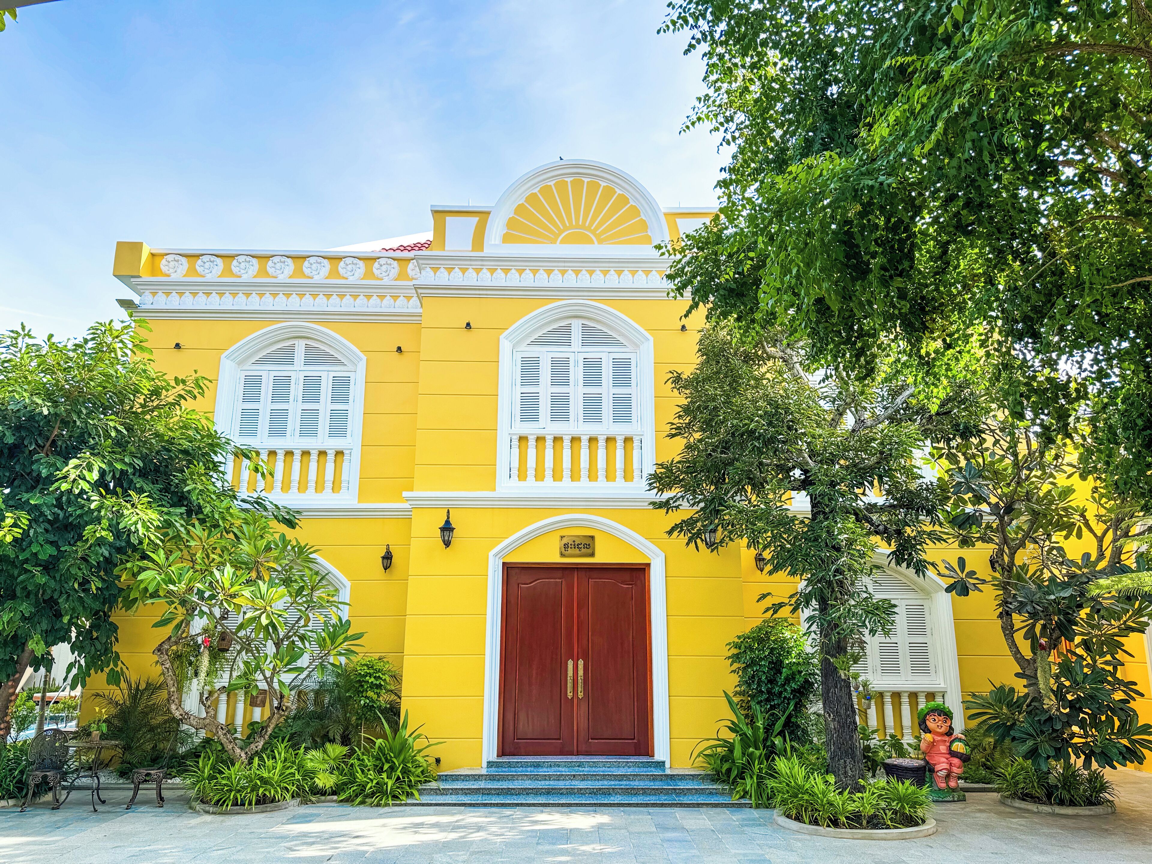 A yellow building with white window shutters, a red door, and a small garden with plants and a statue.