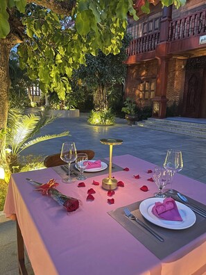 An outdoor dining setup with a table set for two, surrounded by greenery and a brick building. - Piseth Private Villa Hotel (Battambang)