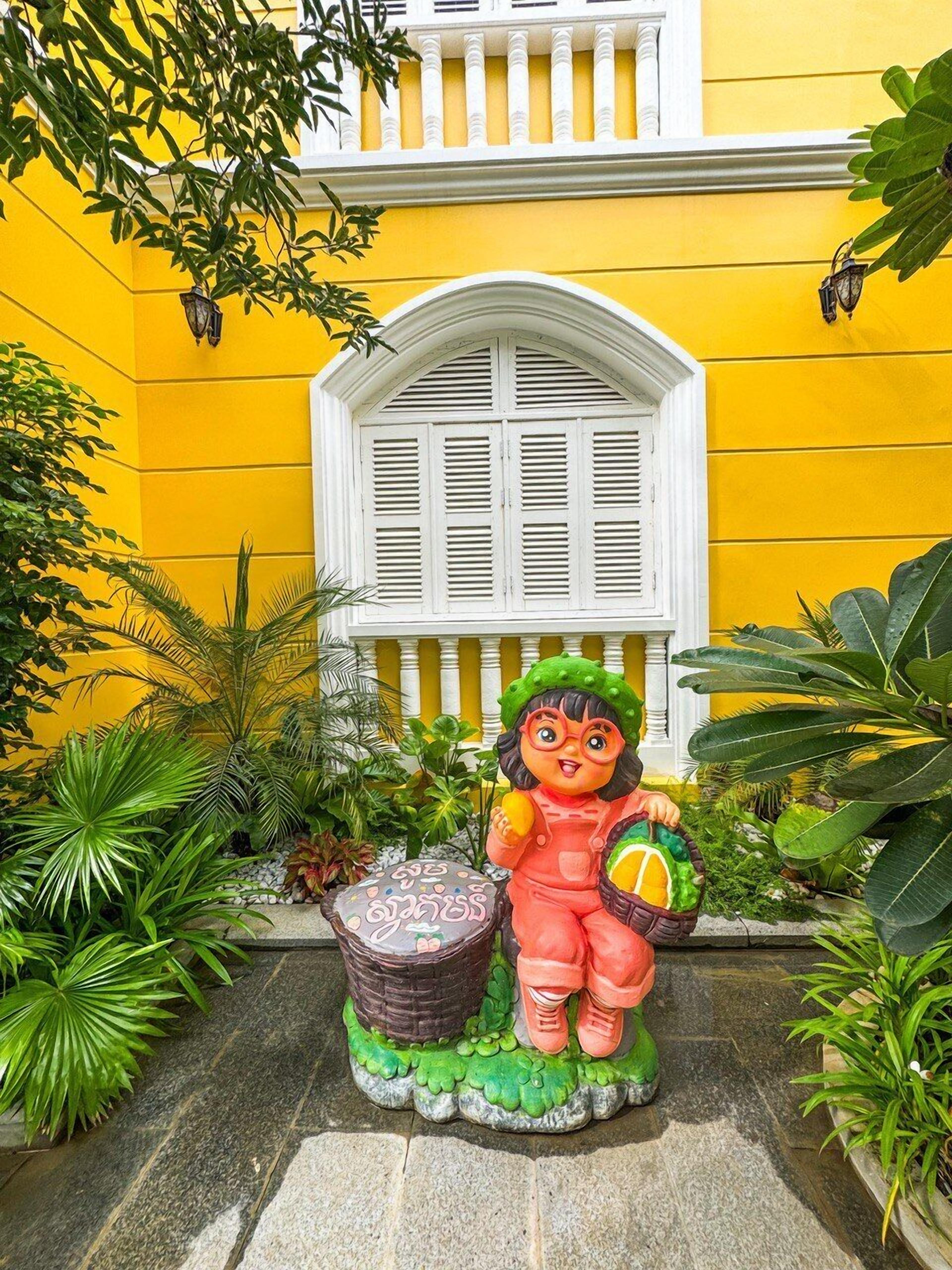 A statue of a child holding a basket of fruit in front of a yellow building with a white window and balcony.
