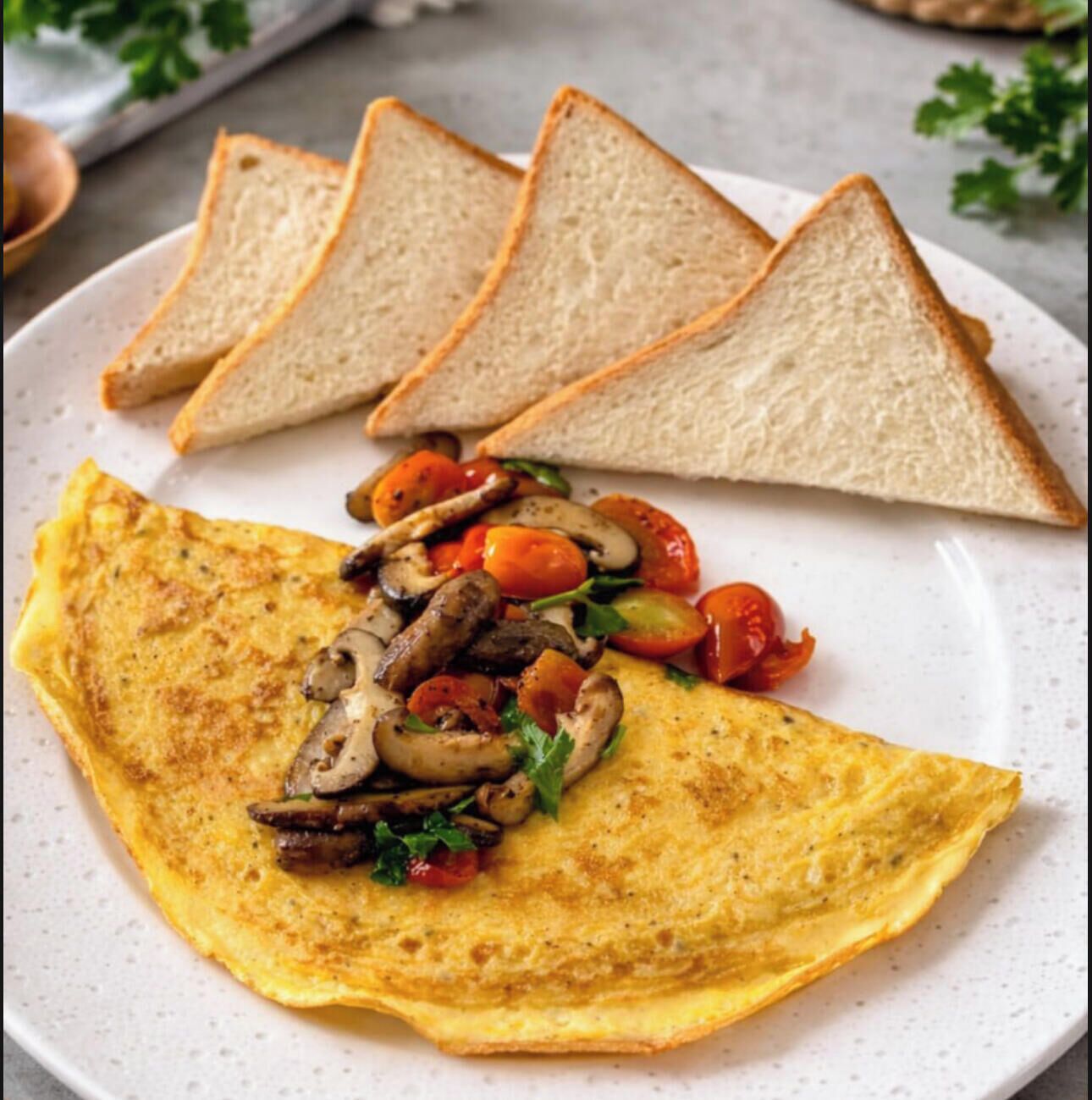 Une assiette contenant une omelette, des champignons en tranches, des tomates et du persil, accompagnée de quatre tranches de pain grillé.
