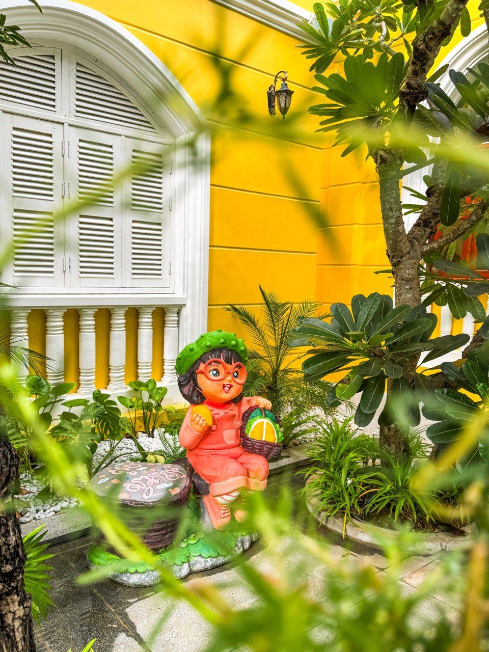 Une statue d’un enfant tenant un panier et un fruit, placée dans un jardin où poussent des plantes tropicales, avec un bâtiment jaune en arrière-plan.