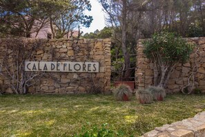 A stone wall with a sign reading 'CALA DE FLORES' surrounded by greenery and trees. - GuestHost - Cala Flores Sea View Apt 400m from the beach! (Porto Cervo)
