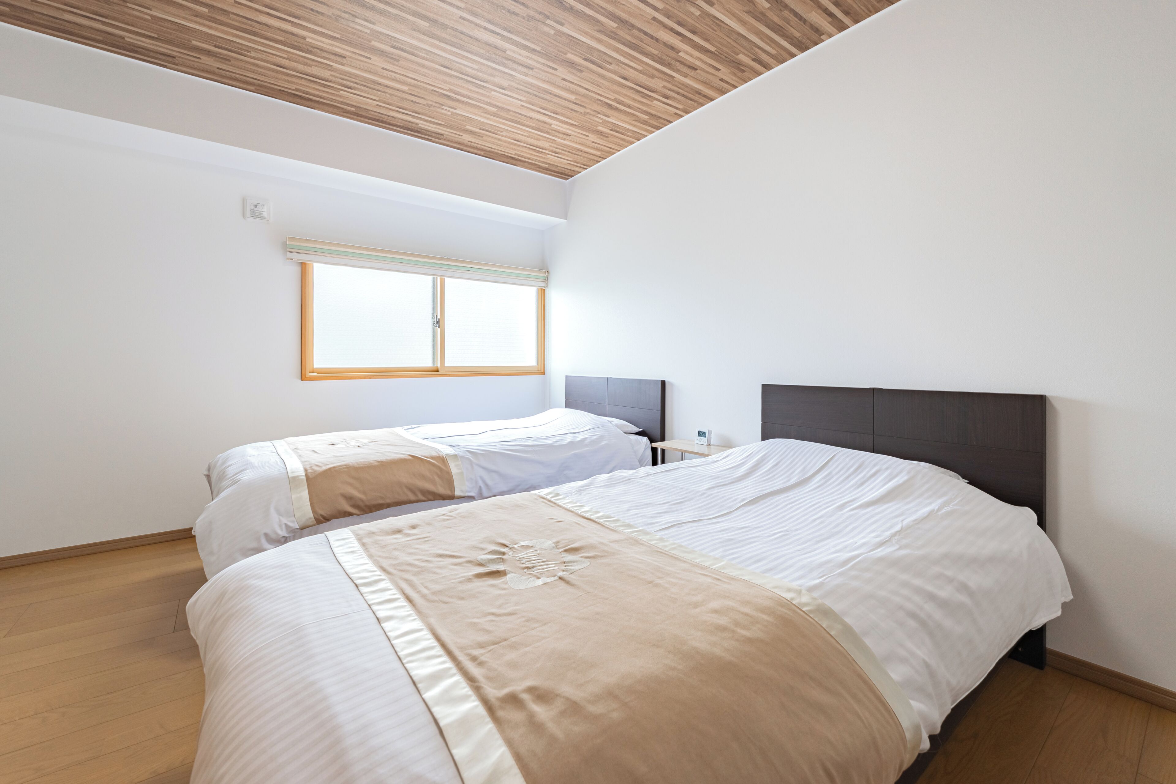 A bedroom with two beds, a wooden ceiling, a window, and a bedside table.