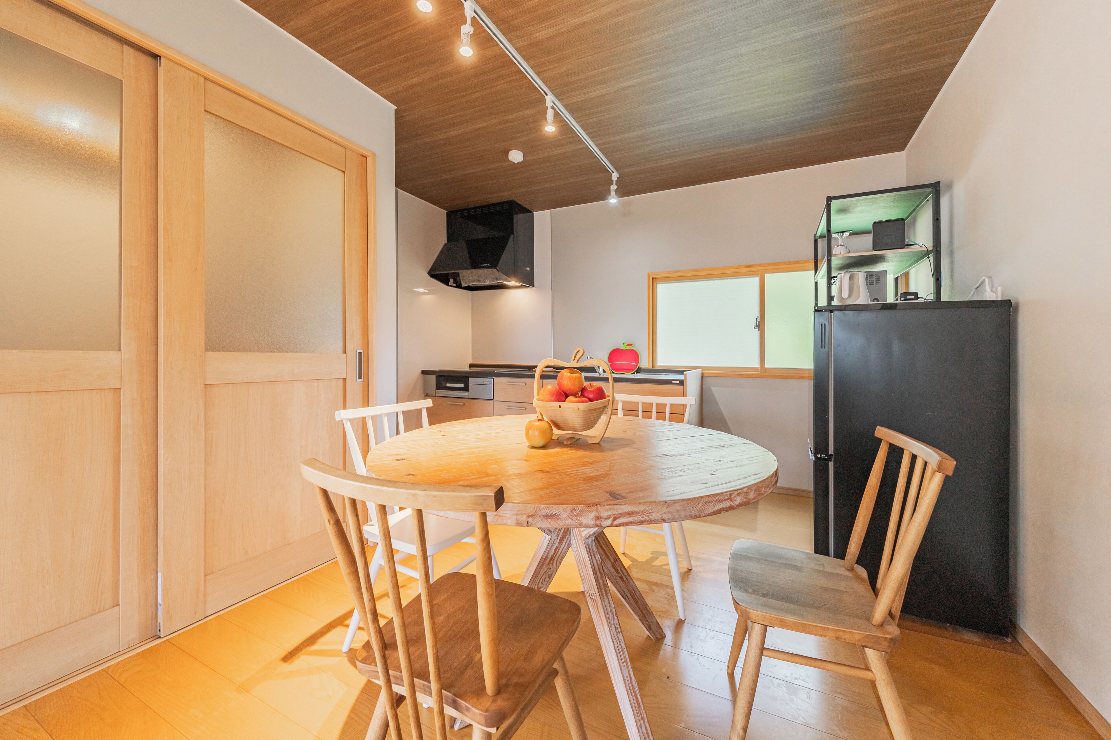 A dining area with a round wooden table, white chairs, and a fruit basket.