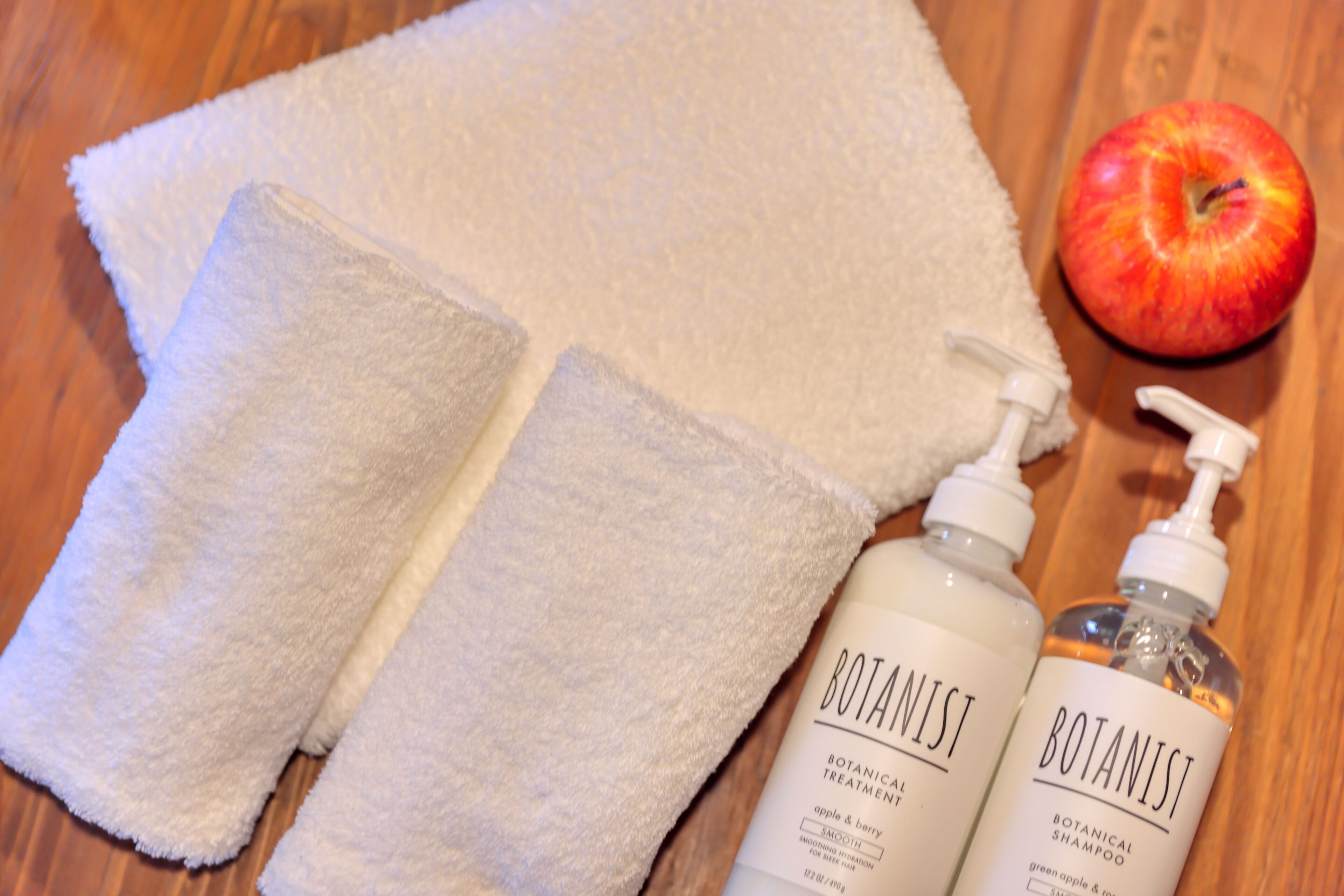 Une serviette blanche, deux bouteilles de shampoing botanique de la marque Botanist, et une pomme sur une surface en bois.