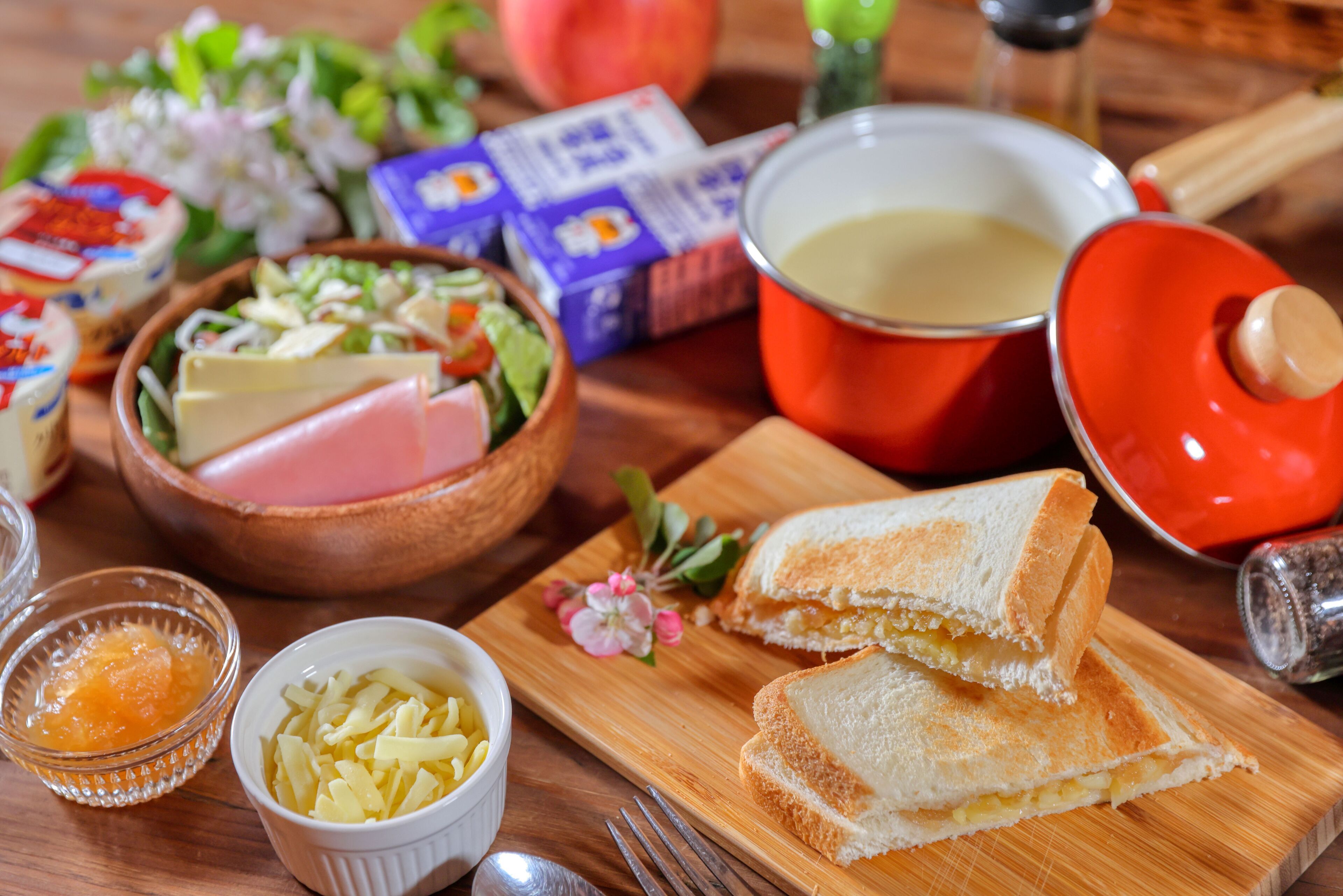 A wooden table with a sandwich, a bowl of salad, a bowl of cheese, a bowl of mustard, and a pot of soup.