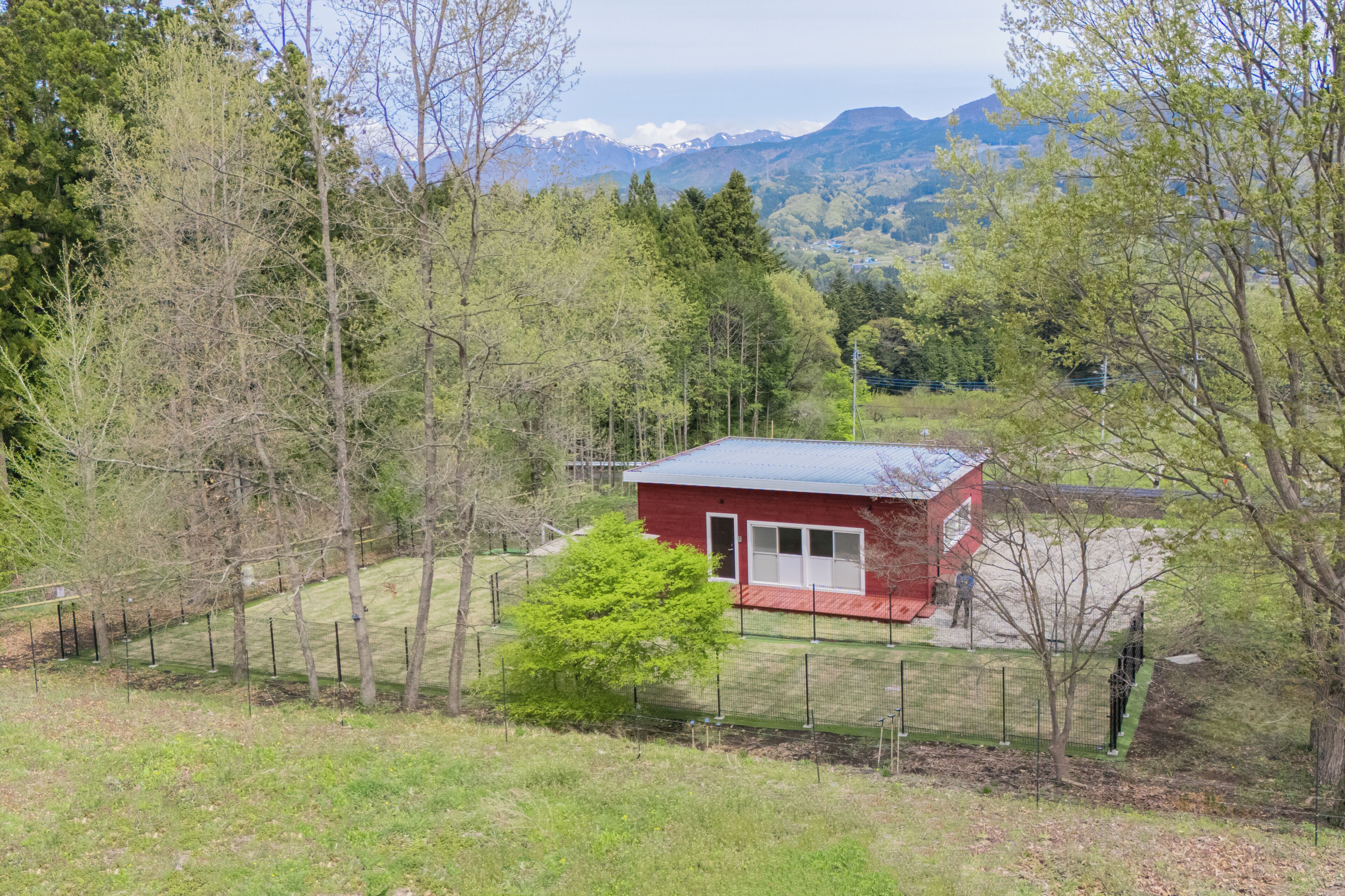 A red building with a fenced yard, surrounded by trees and mountains in the background.