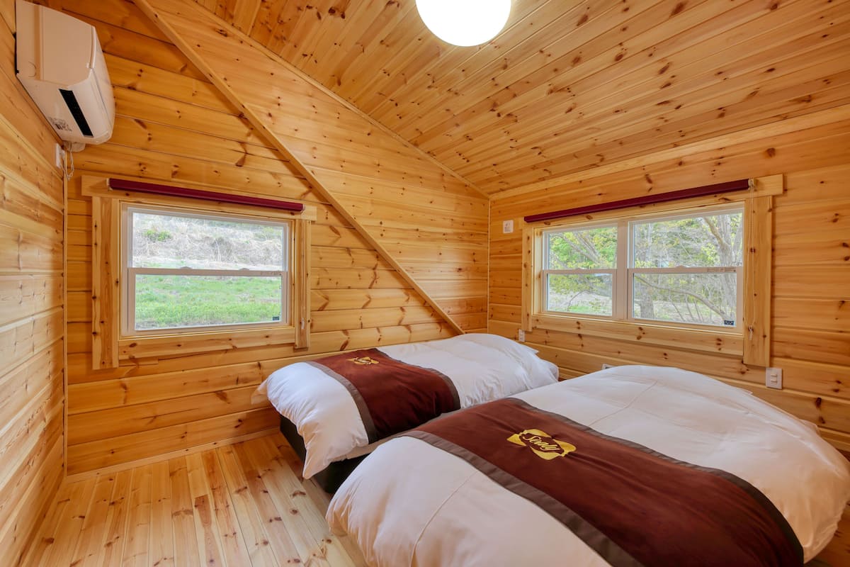 A wooden cabin room with two beds, large windows, and a sloped ceiling.