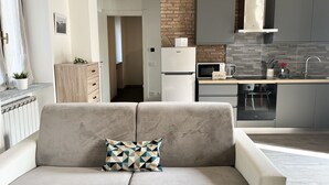Una sala moderna con un sofá y un cojín geométrico, una cocina con armarios grises y una pared de ladrillo decorativa, y vista hacia el pasillo.