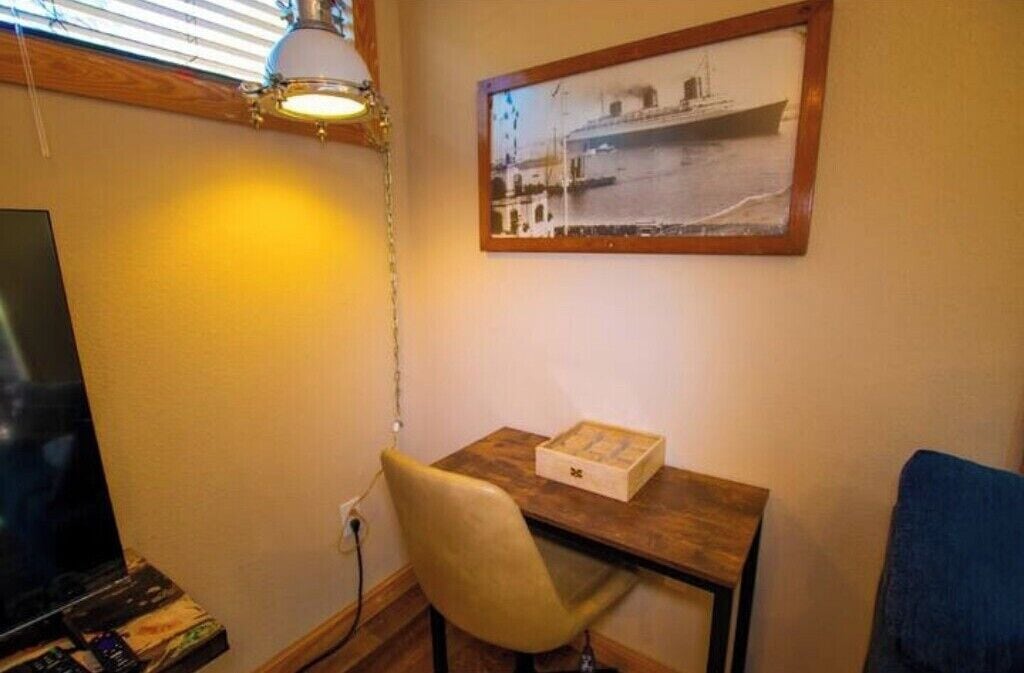 Une petite pièce avec un bureau en bois, une chaise, un tableau encadré au mur et un téléviseur sur le côté gauche.