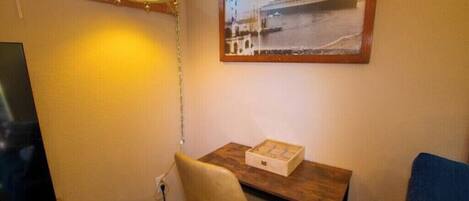 Une petite pièce avec un bureau en bois, une chaise, un tableau encadré au mur et un téléviseur sur le côté gauche.